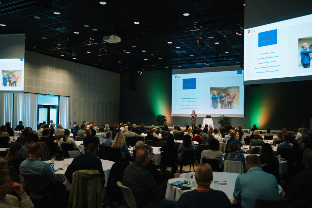 Foto tatt i konferansesal, med publikum, foredragsholder og skjermer.